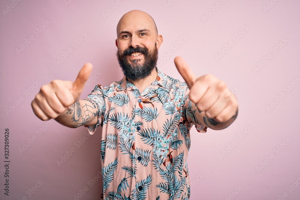 Fototapeta premium Handsome bald man with beard and tattoo wearing casual floral shirt over pink background approving doing positive gesture with hand, thumbs up smiling and happy for success. Winner gesture.