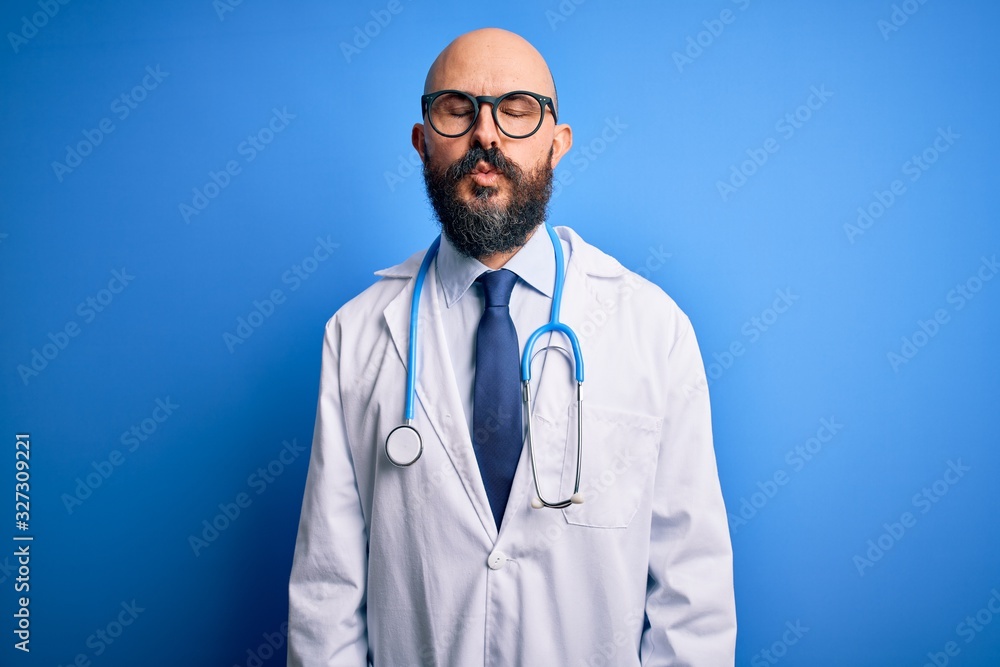 Handsome bald doctor man with beard wearing glasses and stethoscope over blue background looking at the camera blowing a kiss on air being lovely and sexy. Love expression. Handsome bald doctor man with beard wearing glasses and stethoscope over blue background looking at the camera blowing a kiss on air being lovely and sexy. Love expression.