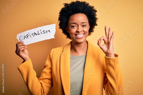 African American afro businesswoman with curly hair holding paper with capitalism message doing ok sign with fingers, excellent symbol photo