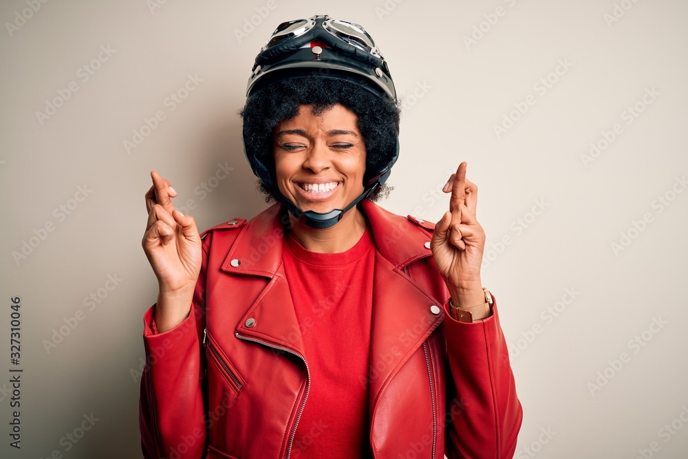 Young African American afro motorcyclist woman with curly hair wearing motorcycle helmet gesturing finger crossed smiling with hope and eyes closed. Luck and superstitious concept. Young African American afro motorcyclist woman with curly hair wearing motorcycle helmet gesturing finger crossed smiling with hope and eyes closed. Luck and superstitious concept.
