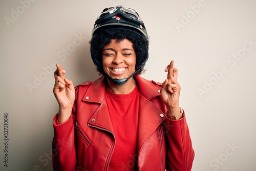Young African American afro motorcyclist woman with curly hair wearing motorcycle helmet gesturing finger crossed smiling with hope and eyes closed. Luck and superstitious concept.