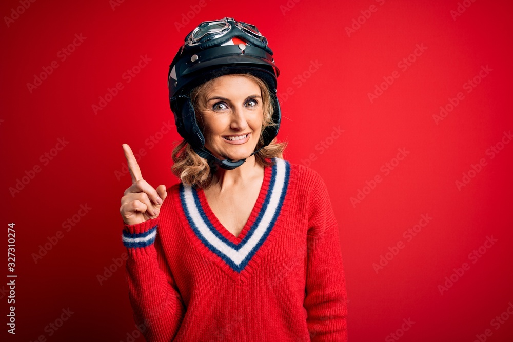Middle age beautiful blonde motorcyclist woman wearing moto helmet over red background pointing finger up with successful idea. Exited and happy. Number one. Middle age beautiful blonde motorcyclist woman wearing moto helmet over red background pointing finger up with successful idea. Exited and happy. Number one.