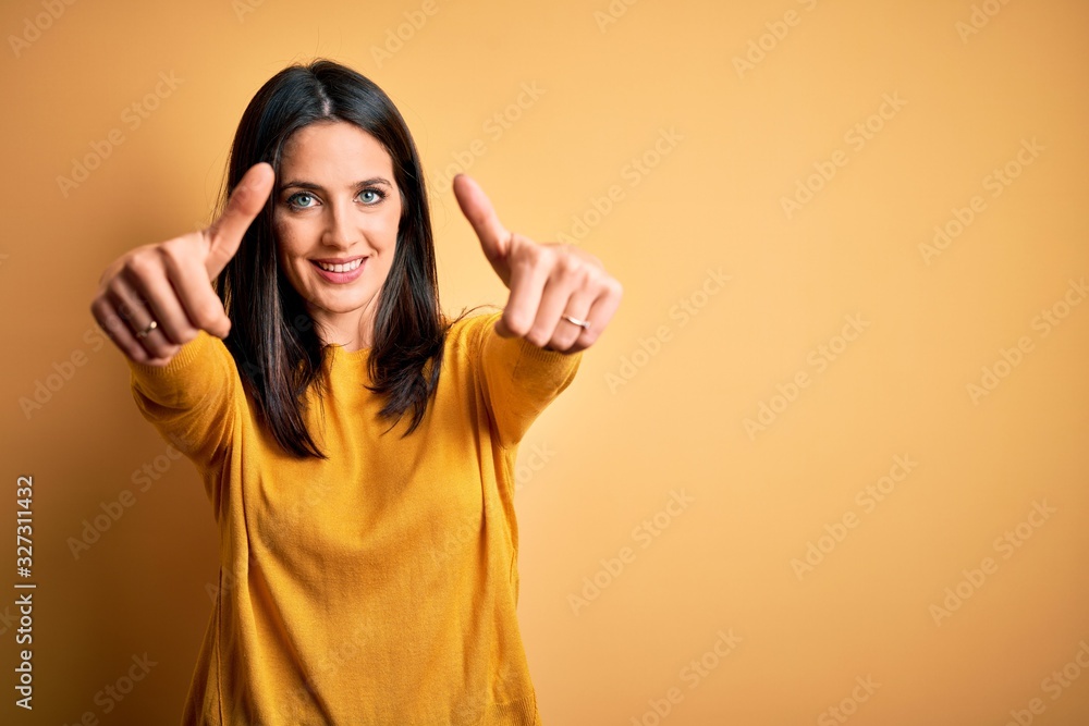 Young brunette woman with blue eyes wearing casual sweater over yellow background approving doing positive gesture with hand, thumbs up smiling and happy for success. Winner gesture. Young brunette woman with blue eyes wearing casual sweater over yellow background approving doing positive gesture with hand, thumbs up smiling and happy for success. Winner gesture.