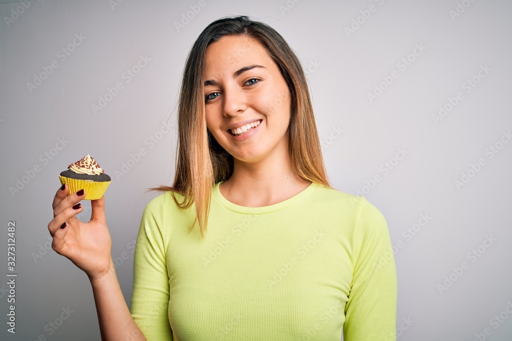 Young beautiful woman with blue eyes eating sweet chocolate cupcake over white background with a happy face standing and smiling with a confident smile showing teeth Young beautiful woman with blue eyes eating sweet chocolate cupcake over white background with a happy face standing and smiling with a confident smile showing teeth