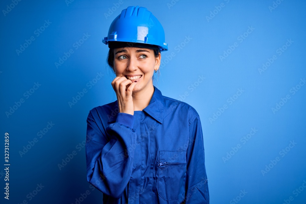 Young beautiful worker woman with blue eyes wearing security helmet and uniform looking stressed and nervous with hands on mouth biting nails. Anxiety problem.