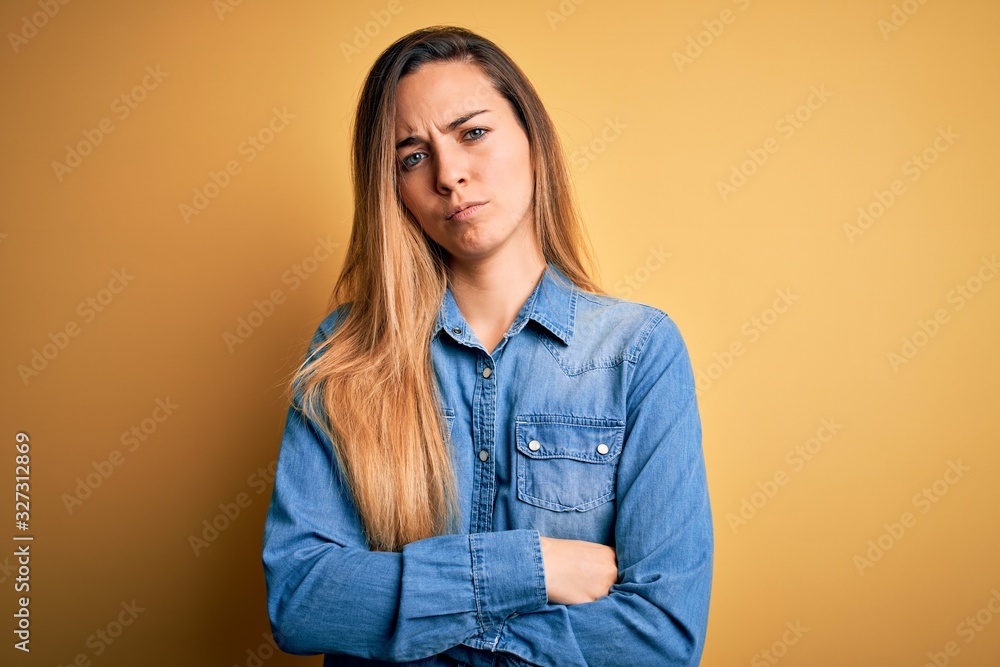 Fototapeta premium Young beautiful blonde woman with blue eyes wearing denim shirt over yellow background skeptic and nervous, disapproving expression on face with crossed arms. Negative person.