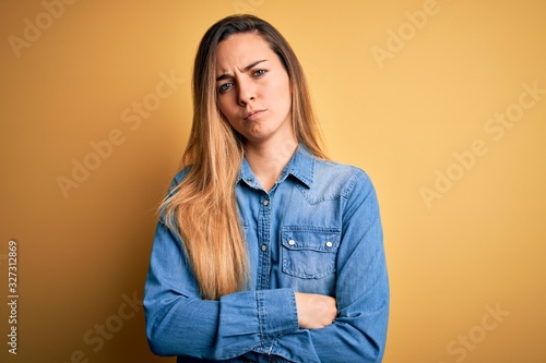 Young beautiful blonde woman with blue eyes wearing denim shirt over yellow background skeptic and nervous, disapproving expression on face with crossed arms. Negative person. © Krakenimages.com
