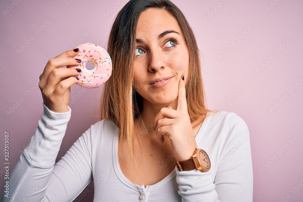Young beautiful blonde woman with blue eyes holding pink doughnut over isolated background serious face thinking about question, very confused idea Young beautiful blonde woman with blue eyes holding pink doughnut over isolated background serious face thinking about question, very confused idea