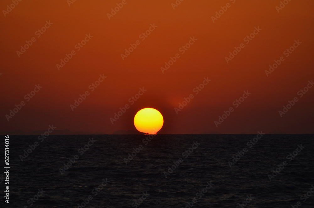 The golden orb of the setting sun has just touched a sea which is almost black. The sky is a deep orange, with dark clouds near the horizon. A few islands are visible on the horizon.