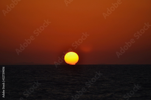 The golden orb of the setting sun has just touched a sea which is almost black. The sky is a deep orange, with dark clouds near the horizon. A few islands are visible on the horizon.