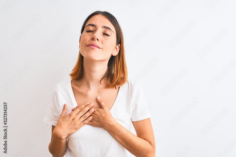 Beautiful redhead woman wearing casual white t-shirt over isolated background smiling with hands on chest with closed eyes and grateful gesture on face. Health concept. Beautiful redhead woman wearing casual white t-shirt over isolated background smiling with hands on chest with closed eyes and grateful gesture on face. Health concept.