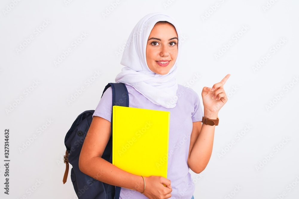 Arab student girl wearing hijab and backpack holding book over isolated white background very happy pointing with hand and finger to the side Arab student girl wearing hijab and backpack holding book over isolated white background very happy pointing with hand and finger to the side