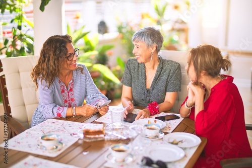 Meeting of middle age women having lunch and drinking coffee. Mature friends smiling happy using smartphone at home on a sunny day