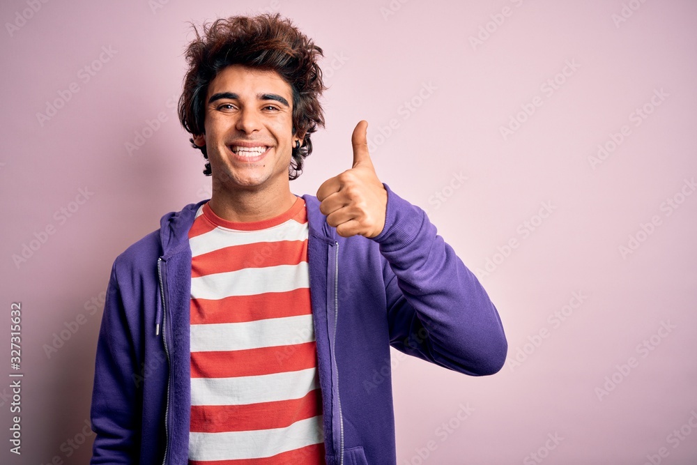 Young man wearing striped t-shirt and purple sweashirt over isolated pink background doing happy thumbs up gesture with hand. Approving expression looking at the camera showing success. Young man wearing striped t-shirt and purple sweashirt over isolated pink background doing happy thumbs up gesture with hand. Approving expression looking at the camera showing success.
