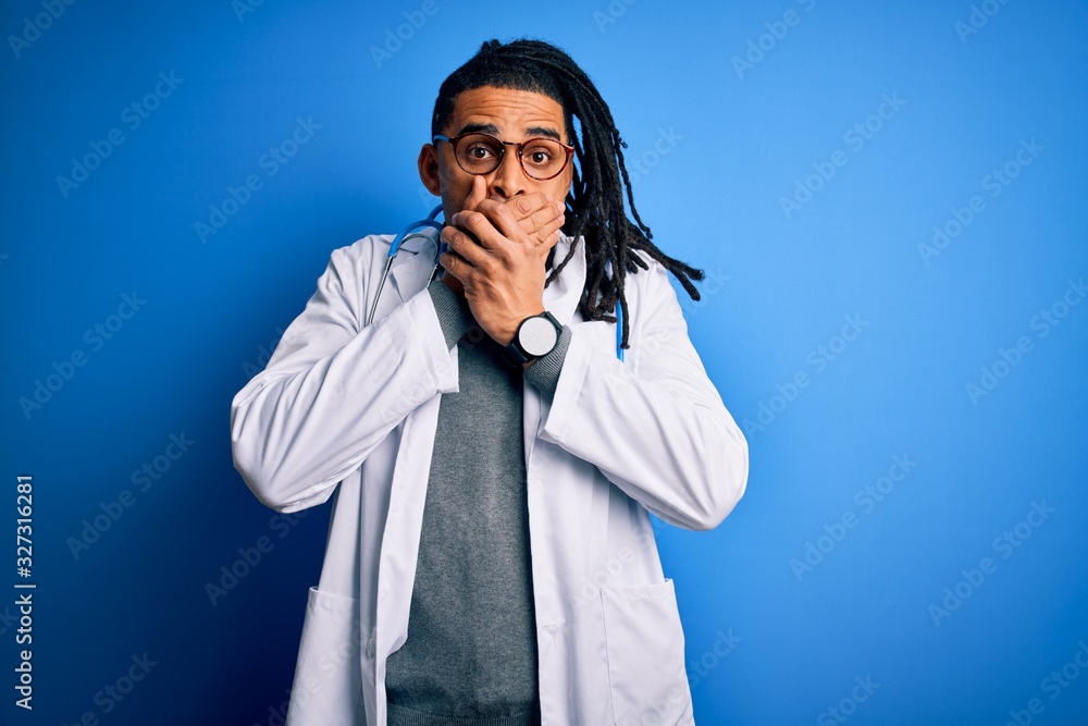 Young african american doctor man with dreadlocks wearing stethoscope and glasses shocked covering mouth with hands for mistake. Secret concept.