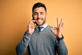 Young handsome man having conversation talking on the smartphone over yellow background doing ok sign with fingers, excellent symbol