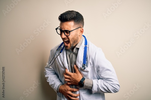 Young doctor man wearing glasses, medical white robe and stethoscope over isolated background with hand on stomach because nausea, painful disease feeling unwell. Ache concept.