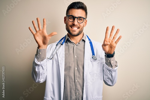 Young doctor man wearing glasses, medical white robe and stethoscope over isolated background showing and pointing up with fingers number nine while smiling confident and happy. © Krakenimages.com