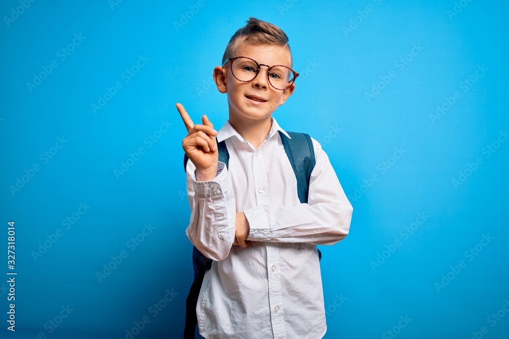 Young little caucasian student kid wearing smart glasses and school bag over blue background with a big smile on face, pointing with hand and finger to the side looking at the camera. Young little caucasian student kid wearing smart glasses and school bag over blue background with a big smile on face, pointing with hand and finger to the side looking at the camera.