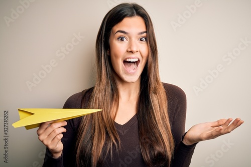 Young beautiful girl holding paper plane standing over isolated white background very happy and excited, winner expression celebrating victory screaming with big smile and raised hands