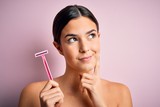 Young beautiful girl using shaver for depilation standing over isolated pink background serious face thinking about question, very confused idea
