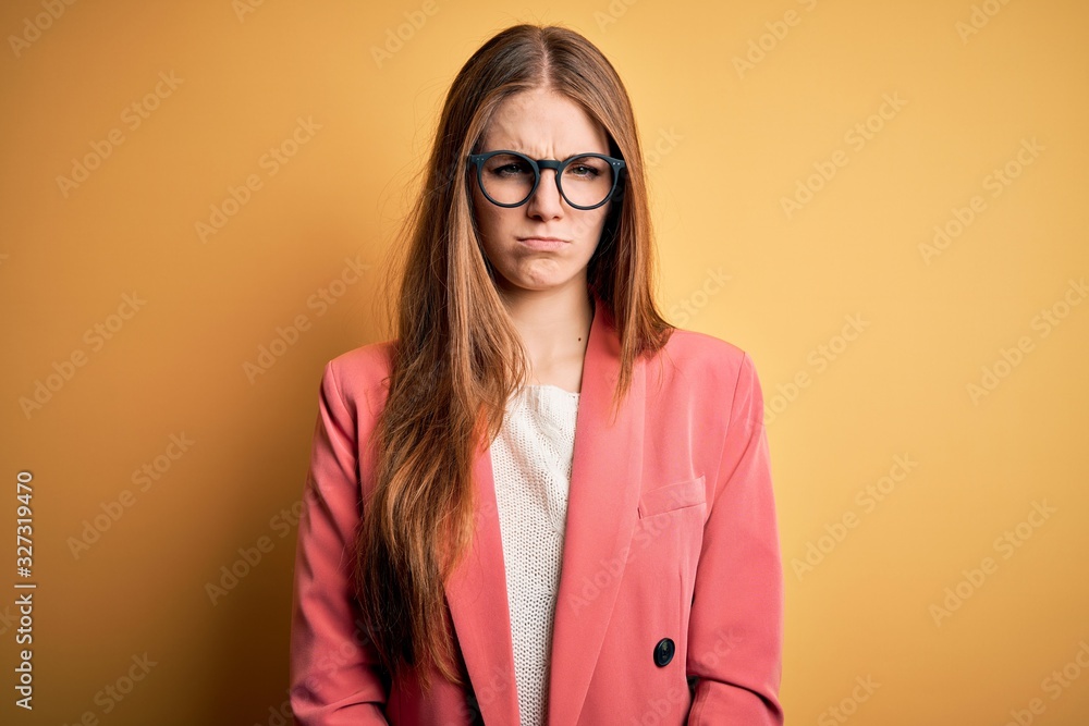 Young beautiful redhead woman wearing jacket and glasses over isolated yellow background skeptic and nervous, frowning upset because of problem. Negative person.