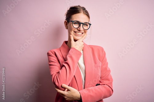 Young beautiful redhead woman wearing jacket and glasses over isolated pink background looking confident at the camera smiling with crossed arms and hand raised on chin. Thinking positive.