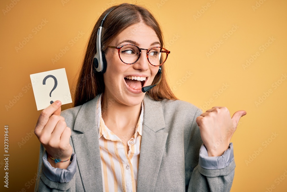 Young redhead call center agent woman using headset holding question mark reminder pointing and showing with thumb up to the side with happy face smiling Young redhead call center agent woman using headset holding question mark reminder pointing and showing with thumb up to the side with happy face smiling