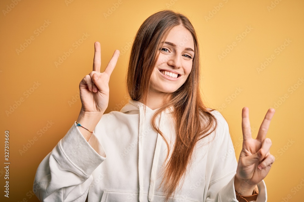 Young beautiful redhead sporty woman wearing sweatshirt over isolated yellow background smiling looking to the camera showing fingers doing victory sign. Number two. Young beautiful redhead sporty woman wearing sweatshirt over isolated yellow background smiling looking to the camera showing fingers doing victory sign. Number two.
