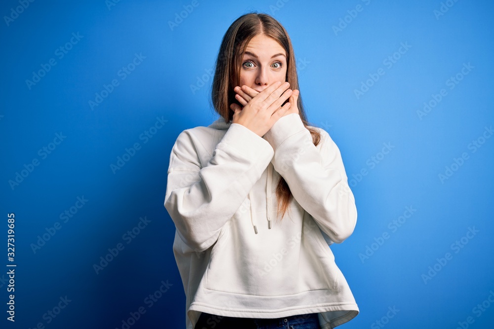 Young beautiful redhead sporty woman wearing sweatshirt over isolated blue background shocked covering mouth with hands for mistake. Secret concept. Young beautiful redhead sporty woman wearing sweatshirt over isolated blue background shocked covering mouth with hands for mistake. Secret concept.