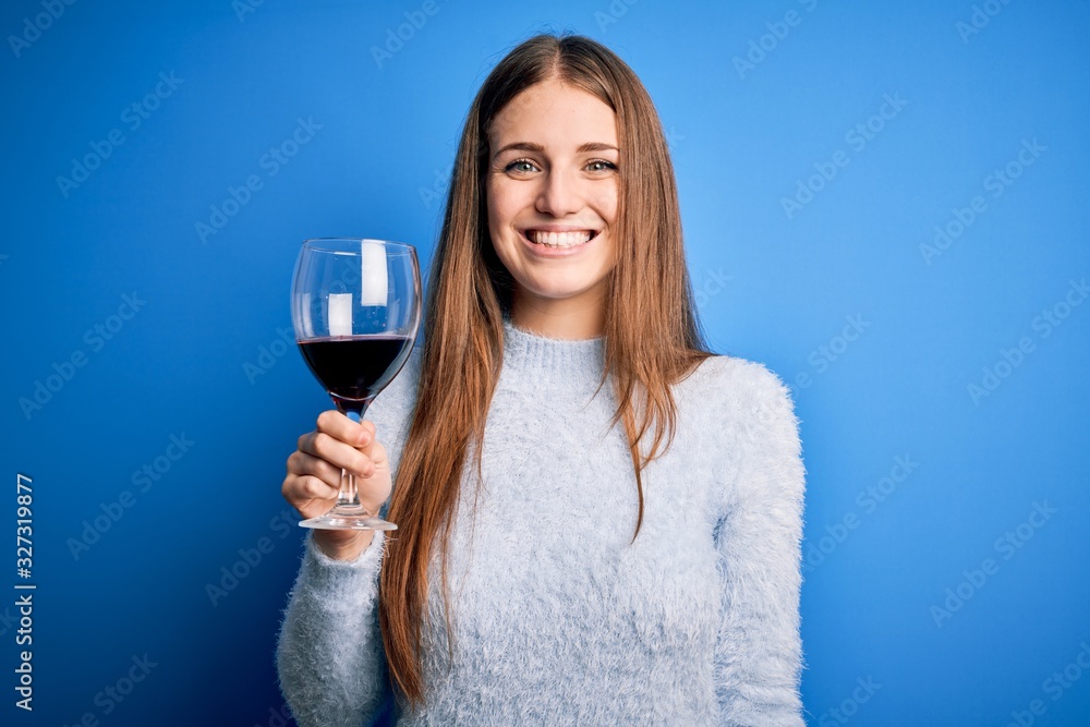 Young beautiful redhead woman drinking glass of red wine over isolated blue background with a happy face standing and smiling with a confident smile showing teeth Young beautiful redhead woman drinking glass of red wine over isolated blue background with a happy face standing and smiling with a confident smile showing teeth