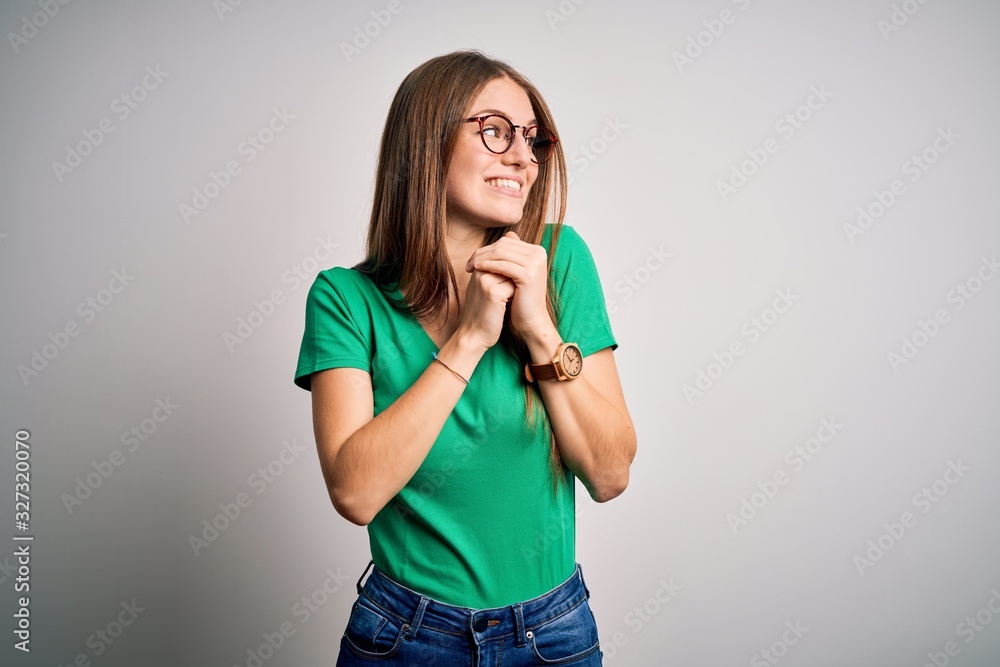 Young beautiful redhead woman wearing casual green t-shirt and glasses over white background laughing nervous and excited with hands on chin looking to the side Young beautiful redhead woman wearing casual green t-shirt and glasses over white background laughing nervous and excited with hands on chin looking to the side