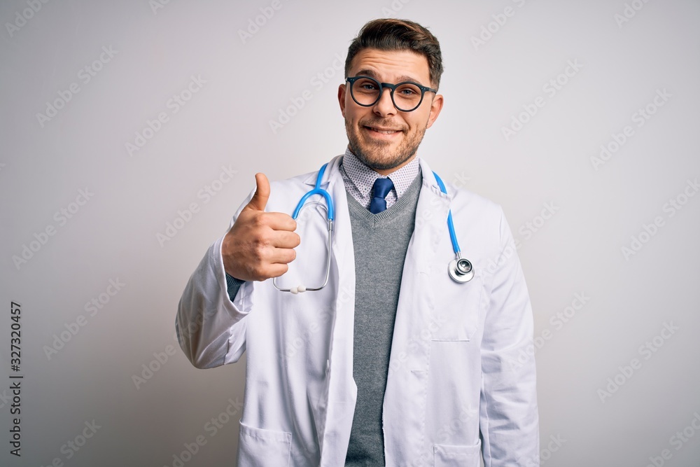 Young doctor man with blue eyes wearing medical coat and stethoscope over isolated background doing happy thumbs up gesture with hand. Approving expression looking at the camera showing success. Young doctor man with blue eyes wearing medical coat and stethoscope over isolated background doing happy thumbs up gesture with hand. Approving expression looking at the camera showing success.
