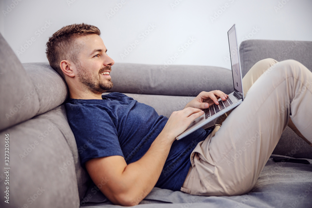 Side view of attractive bearded smiling man lying on sofa and using laptop for internet surfing. - obrazy, fototapety, plakaty 