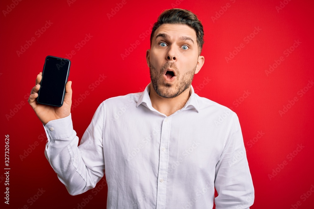 Young business man with blue eyes showing smartphone screen over red background scared in shock with a surprise face, afraid and excited with fear expression Young business man with blue eyes showing smartphone screen over red background scared in shock with a surprise face, afraid and excited with fear expression
