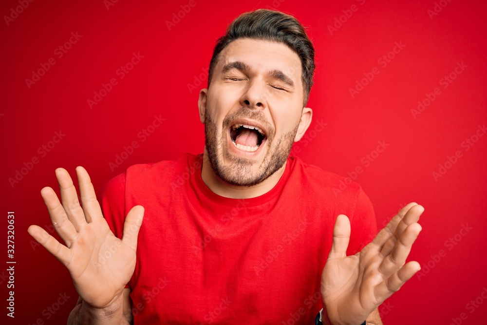 Young man with blue eyes wearing casual t-shirt over red isolated background celebrating mad and crazy for success with arms raised and closed eyes screaming excited. Winner concept Young man with blue eyes wearing casual t-shirt over red isolated background celebrating mad and crazy for success with arms raised and closed eyes screaming excited. Winner concept