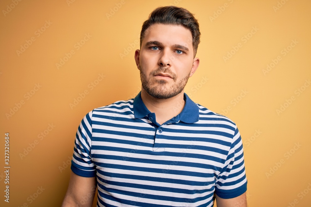 Young man with blue eyes wearing casual striped t-shirt over yellow background with serious expression on face. Simple and natural looking at the camera. Young man with blue eyes wearing casual striped t-shirt over yellow background with serious expression on face. Simple and natural looking at the camera.