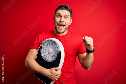 Young fitness man with blue eyes holding scale dieting for healthy weight over red background screaming proud and celebrating victory and success very excited, cheering emotion