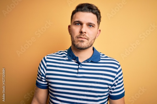 Young man with blue eyes wearing casual striped t-shirt over yellow background with serious expression on face. Simple and natural looking at the camera.