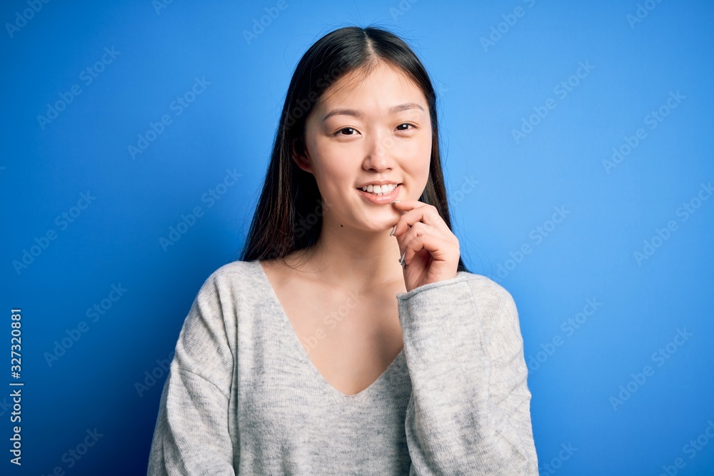 Young beautiful asian woman wearing casual sweater standing over blue isolated background looking confident at the camera smiling with crossed arms and hand raised on chin. Thinking positive. Young beautiful asian woman wearing casual sweater standing over blue isolated background looking confident at the camera smiling with crossed arms and hand raised on chin. Thinking positive.