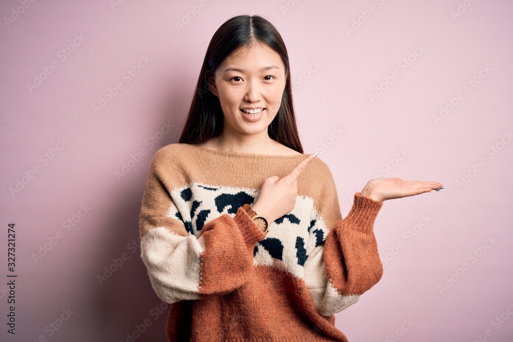 Young beautiful asian woman wearing animal print fashion sweater over pink isolated background amazed and smiling to the camera while presenting with hand and pointing with finger. Young beautiful asian woman wearing animal print fashion sweater over pink isolated background amazed and smiling to the camera while presenting with hand and pointing with finger.