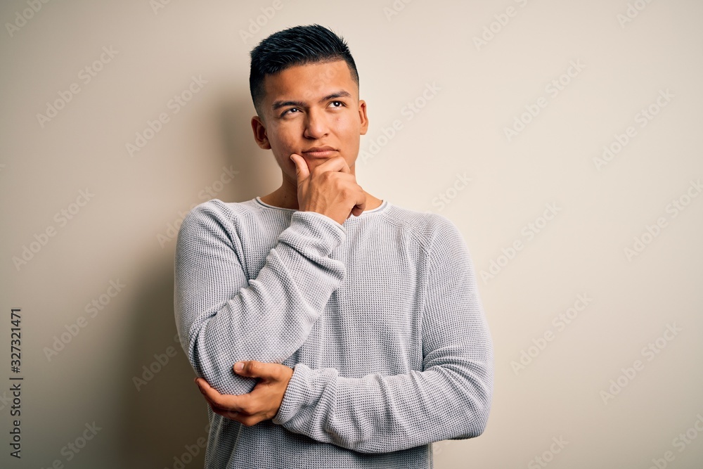 Young handsome latin man wearing casual sweater standing over isolated white background with hand on chin thinking about question, pensive expression. Smiling with thoughtful face. Doubt concept. Young handsome latin man wearing casual sweater standing over isolated white background with hand on chin thinking about question, pensive expression. Smiling with thoughtful face. Doubt concept.
