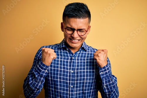 Young handsome latin man wearing casual shirt and glasses over yellow background excited for success with arms raised and eyes closed celebrating victory smiling. Winner concept.