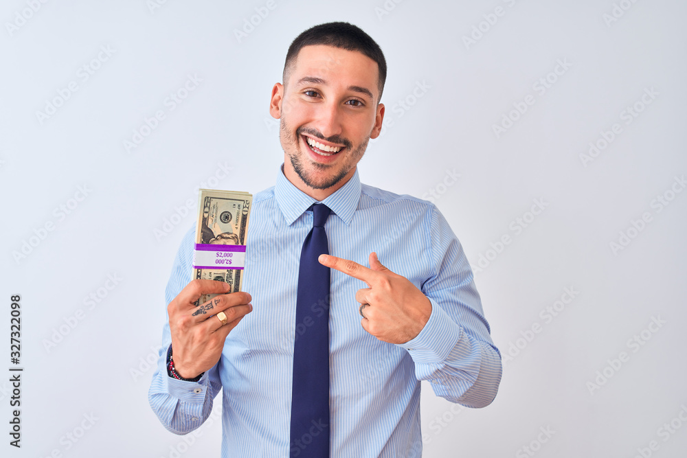 Young handsome business man holding a bunch of dollars bank notes over isolated background very happy pointing with hand and finger Young handsome business man holding a bunch of dollars bank notes over isolated background very happy pointing with hand and finger