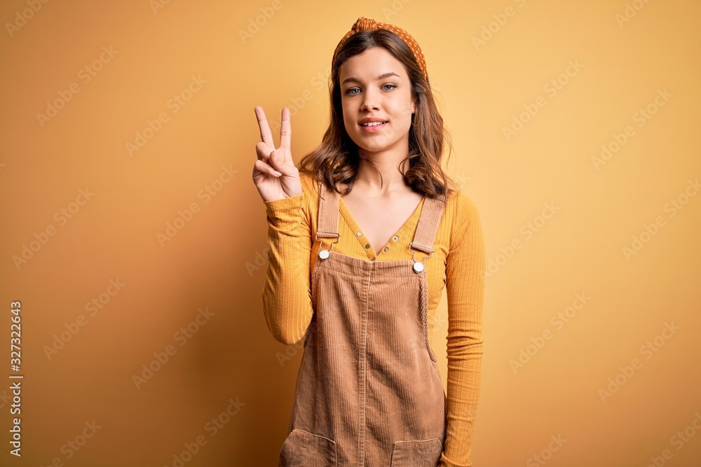 Young beautiful blonde girl wearing overall standing over yellow isolated background showing and pointing up with fingers number two while smiling confident and happy. Young beautiful blonde girl wearing overall standing over yellow isolated background showing and pointing up with fingers number two while smiling confident and happy.