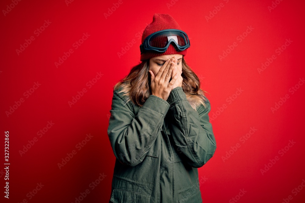 Young blonde girl wearing ski glasses and winter coat for ski weather over red background with sad expression covering face with hands while crying. Depression concept. Young blonde girl wearing ski glasses and winter coat for ski weather over red background with sad expression covering face with hands while crying. Depression concept.