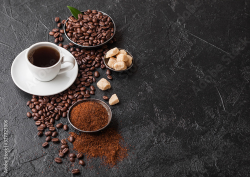 White cup of fresh raw organic coffee with beans and ground powder with cane sugar cubes with coffee tree leaf on black background. Top view. Space for text
