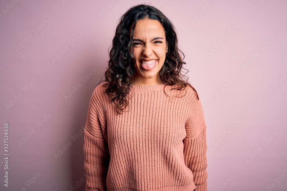 Young beautiful woman with curly hair wearing casual sweater over isolated pink background sticking tongue out happy with funny expression. Emotion concept. Young beautiful woman with curly hair wearing casual sweater over isolated pink background sticking tongue out happy with funny expression. Emotion concept.