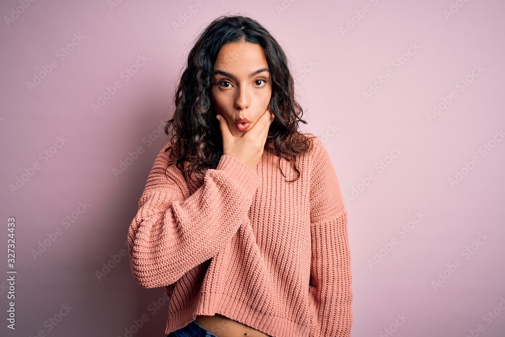 Young beautiful woman with curly hair wearing casual sweater over isolated pink background Looking fascinated with disbelief, surprise and amazed expression with hands on chin Young beautiful woman with curly hair wearing casual sweater over isolated pink background Looking fascinated with disbelief, surprise and amazed expression with hands on chin