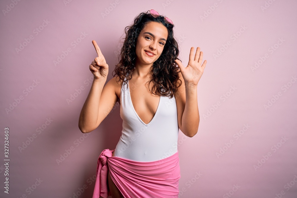Beautiful woman with curly hair on vacation wearing white swimsuit over pink background showing and pointing up with fingers number six while smiling confident and happy. Beautiful woman with curly hair on vacation wearing white swimsuit over pink background showing and pointing up with fingers number six while smiling confident and happy.
